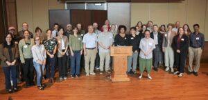 Approximately 30 participants pose for a group photo, standing in several rows behind a wooden podium.