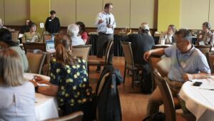 A presenter stands in the center of a meeting room speaking into a microphone while participants seated at round tables listen and engage in discussion.